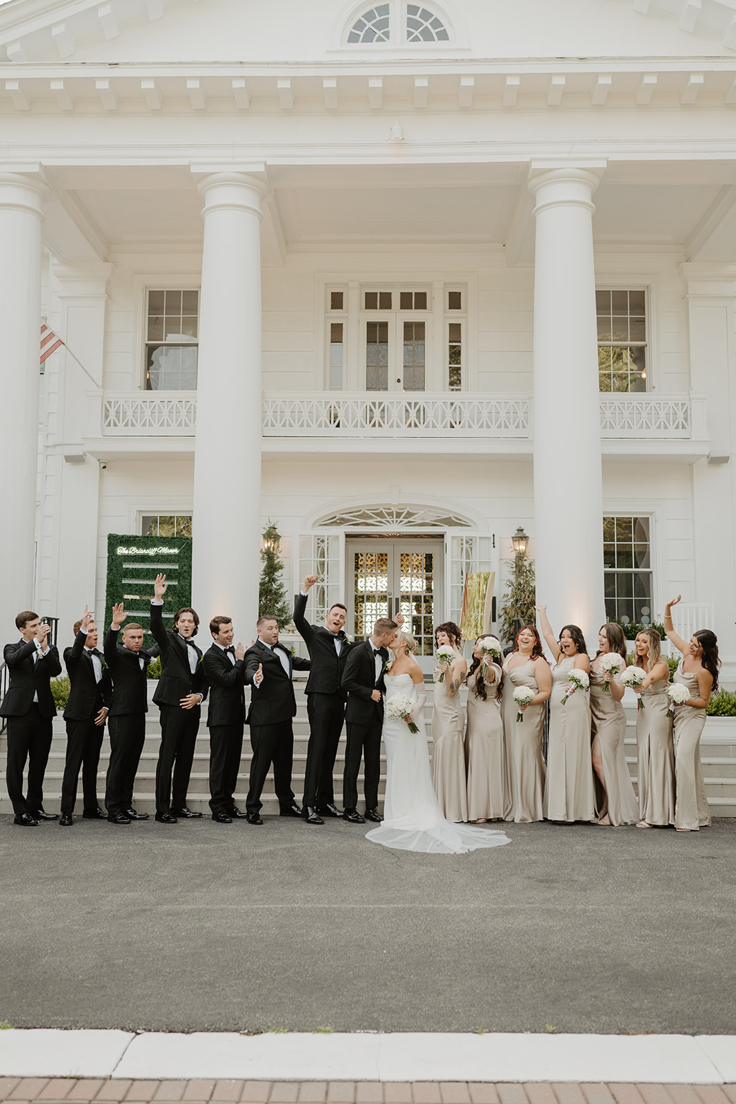 Wedding party cheers on the steps of the grand white manor as the bride and groom kiss in the center.